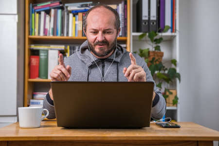 Bearded man working online from home on computer laptop behind vintage desk, with earphones, talking and explaining to colleagues on online meetingの写真素材