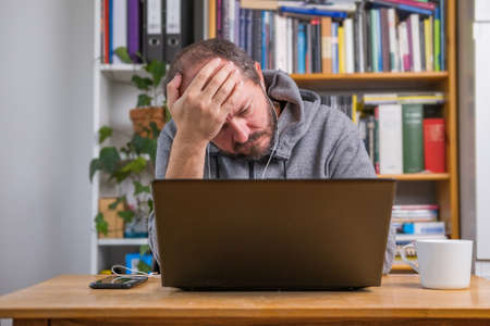 Man working online from home office on computer laptop behind vintage desk, with earphones, tired and exhausted after online meeting, facepalm expressionの写真素材
