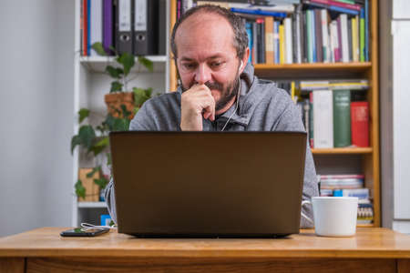 Man working online from home office on computer laptop behind vintage desk, with earphones, listening online meeting and drinking coffeeの写真素材