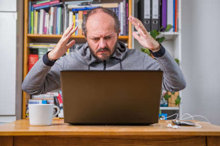 Angry man working online from home office on computer laptop behind vintage desk, arms gesture, business problem conceptの写真素材