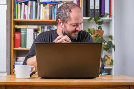 Man working online from home office on computer laptop behind vintage desk, focused, with earphones, listening online meetingの写真素材