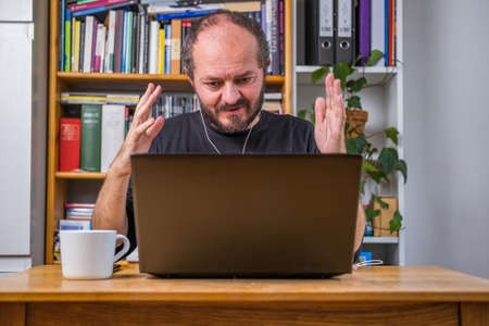 Man working online from home office on computer laptop behind vintage desk, with earphones, listening online meeting and drinking coffeeの写真素材