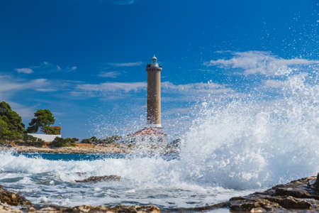 Sea waves breaking on rocks, lighthouse of Veli Rat on the island of Dugi Otok, Croatia in backgroundの写真素材