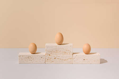 Three eggs standing on a travertine marble podium on a beige and gray background. 2021 Easter unique...