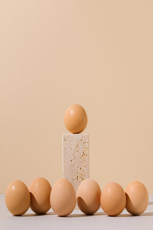 Single egg standing on a travertine marble block stage surrounded by group of eggs on a beige and...