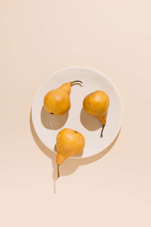 Yellow pears on a white ceramic plate casting shadows against pale beige background. Sunlit summer fruit concept. Creative flat lay arrangement.の写真素材