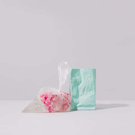 Pink peony flowers in a plastic freezer bag filled with water leaning against blue ceramic vase on a grey background. Surreal art direction floral still life. Abstract rose flower concept.の写真素材