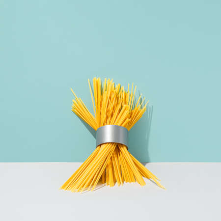 Raw spaghetti and duct tape roll composition against cyan blue background. Abstract yellow pasta with grey adhesive tape creative concept. Surreal italian food art direction.の写真素材