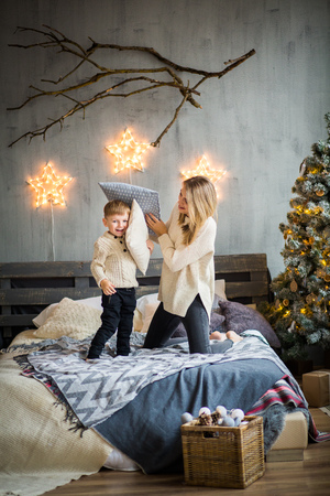 Happy family portrait of mother and baby boy playing with pillows on cosy bed in festivaly decorated room with Christmass tree and shining stars on the wall. New Year`s celebration.の写真素材