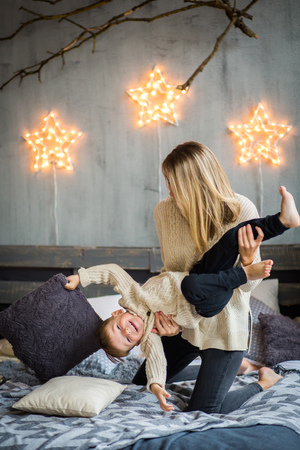 Mother and little boy are having fun on cosy bed in front of festivaly decorated wall with shining stars. Cute boy is laughing. New Year`s celebration.の写真素材