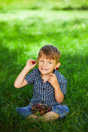 Happy little boy with ripe cherry berries in his hands sittng on green grass on a sunny summer day.の写真素材
