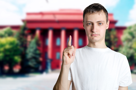 Serious young guy showing finger up against background of university. The man calls for attention, caution. The concept of an important statement, information. Danger, seriousness, severity.の写真素材