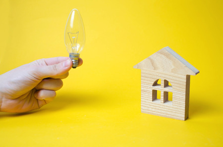 A wooden house and a light bulb in his hand. Concept ideas, protective environment, replacement of light bulbs in the house. Wooden toy on a yellow background. Conservation of energy and innovation.の写真素材