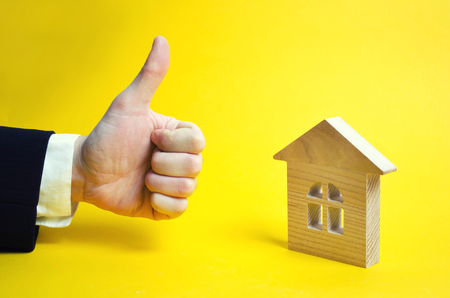 Finger up and house. Wooden house and approving hand. A businessman in a suit gives a good appraisal to the business. Real estate, good make, excellent price. Yellow background.の写真素材