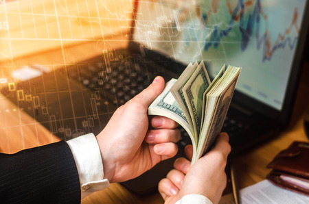 A businessman in a suit counts dollars at a table in the hands of a laptop and financial charts. A concept of a successful business, successful selling, savings, investment. Playing on the exchange.の写真素材
