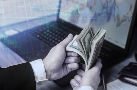A businessman in a suit counts dollars at a table in the hands of a laptop and financial charts. A concept of a successful business, successful selling, savings, investment. Playing on the exchange.の写真素材
