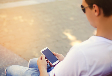 the guy is holding a mobile smartphone and looking at the screen. telephone dependence, social networks. work on the Internet. write message. sunny day. close-up, space for textの写真素材