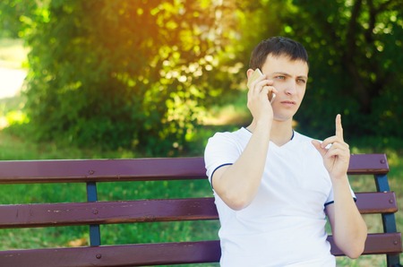 a young European guy in a white T-shirt speaks on the phone and sits on a bench in the city park and points with your finger. The concept of solving problems, unexpected news.の写真素材