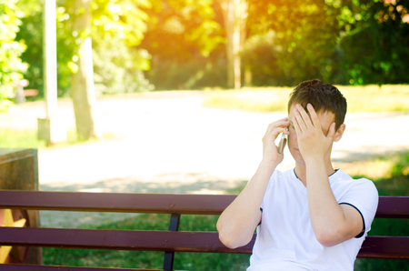 a young pensive European guy in a white T-shirt speaks on the phone and sits on a bench in the city park. covers his face with his hands. the man is upset, the bad unexpected news. The concept of solving problemsの写真素材