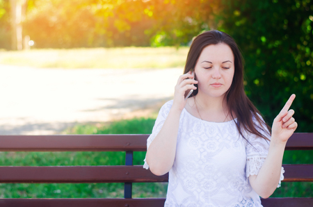 Young beautiful european girl sitting on a bench and talking on the phone. The girl points a finger away, gives advice and directions to the interlocutor by phone. Teachings, advice, judgment.の写真素材