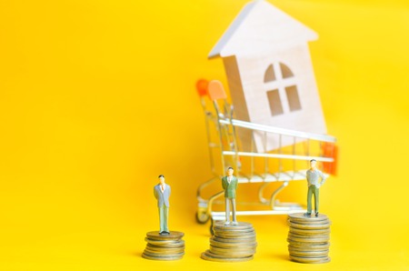 People stand on coins on the background of a wooden house and a basket from a supermarket. Concept of real estate. Buying, selling and renting a house. Loan for an apartment, mortgage rate. Affordable housing for young families.の写真素材