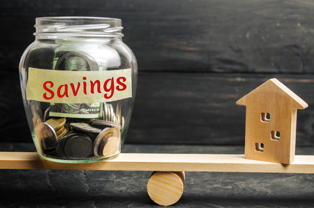 Miniature house and glass jar with coins on the scales. The inscription "savings". The accumulation of money to buy a house, apartment, real estate. Purchase a property.の写真素材