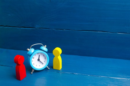 Two men stand near the blue alarm clock and talk. The concept of waiting for a meeting, a date. Punctuality. The cost of hourly work of a specialist, business planning. Discussing cases.の写真素材