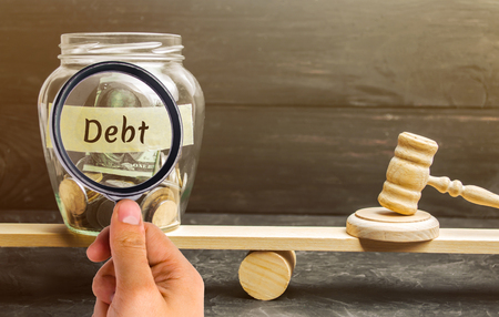 A glass jar with coins and the inscription "Debt" and the judge's hammer are on the scales. Concept of financial crisis and problems. Risk management concept. Payment of taxes. Debt exemption. Court.の写真素材