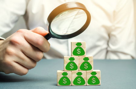 Businessman holds magnifying glass over cubes with dollars. Analysis of profits and revenues in the company. Distribution of money. The accumulation of capital. Budget planning. Investmentsの写真素材