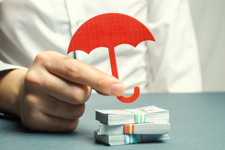 An insurance agent holds a red umbrella over dollar bills. Savings protection. Keeping money safe. Investment and capital insurance. The risk of doing business. Corruption losses. Arrest of investorsの写真素材