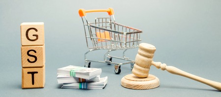 Wooden blocks with the word GST, money and a supermarket trolley with a judge's gavel. Tax, which is imposed on the sale of goods and servicesの写真素材