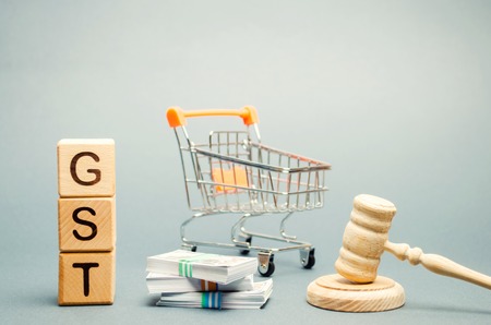 Wooden blocks with the word GST, money and a supermarket trolley with a judge's gavel. Tax, which is imposed on the sale of goods and servicesの写真素材