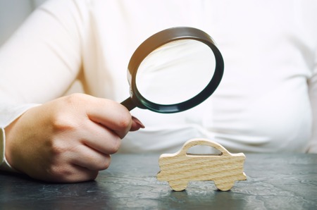A woman holds a magnifying glass over a miniature wooden car. The estimated cost of the auto. Analysis and technical inspection of cars. Buying a new car. Insurance. Search. Vehicle condition. Loan.の写真素材