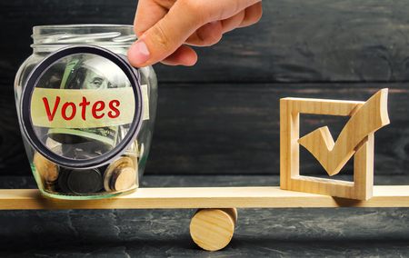 Glass jar with coins and the words "Votes" and a checkbox on the scales. Concept of voting for money. Bribing voters. Corruption in the electoral system. Interference in the election. Lobbying.の写真素材
