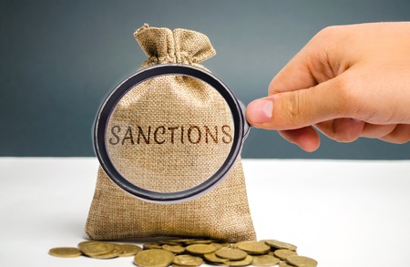 A magnifying glass is looking at a money bag with the word Sanctions. The imposition of economic and political sanctions on the subjects of geopolitics. Restriction and pressure of the world communityの写真素材