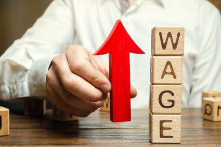 Businessman's hand holds red arrow up near wooden blocks with word Wage. Salary increase concept. Wages rate. Revenue growth and profits. Successful business. Career rise. Promote. Stabilityの写真素材