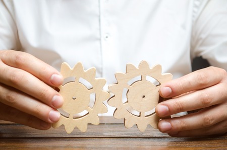 Businessman in white shirt connects two wooden gears. Symbolism of establishing business processes and communication. Improving work efficiency, establishing new connections and suppliers.の写真素材
