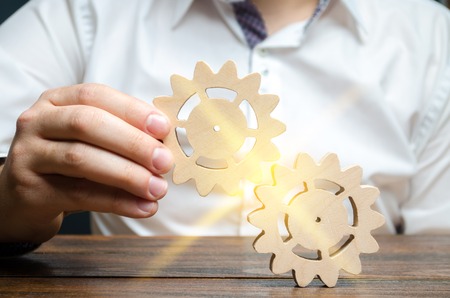 Businessman in white shirt connects two wooden gears. Symbolism of establishing business processes and communication. Increase efficiency and productivity. The best business formula for success.の写真素材
