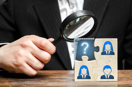 Businessman with a magnifying glass examines the missing part of the puzzle team. Search, recruitment staff, hiring leader. Creating an effective business team. Lack of specialists in the labor marketの写真素材