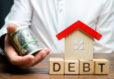 Wooden blocks with the word Debt, house and dollars in the hands of a businessman. The concept of timely payment of debt for real estate. Buying a property in debt. Mortgage. Credit for the apartment.の写真素材