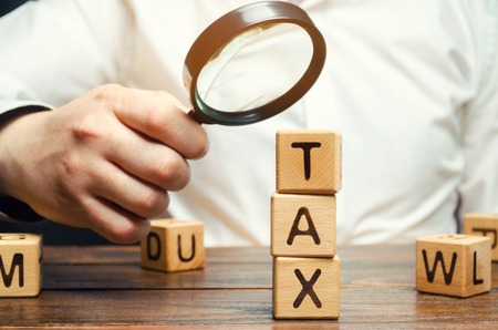 Wooden blocks with the word Tax and a magnifying glass in the hands of a businessman. The concept of studying the size of taxes. Taxation. Time to pay taxes. Tax on personal income. Agent. Taxpayerの写真素材