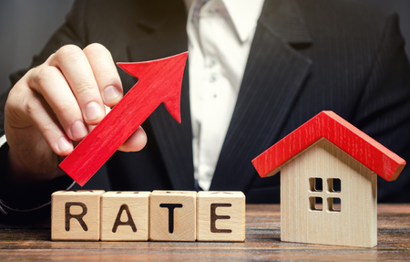A man holds a red arrow up above the word Rate and a wooden house. The concept of raising interest rates on mortgages. The increase in property tax rates. Real estate capitalization. Insurance.の写真素材