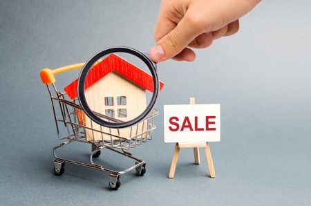 Magnifying glass is looking at the Supermarket cart with houses and a Sale Poster. The concept of selling a home, real estate services or buying from the owner. property, apartments. Selective focusの写真素材