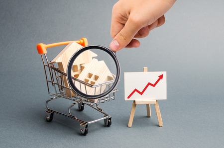 Magnifying glass is looking at the Wooden houses in a supermarket cart and red up arrow. Growing demand for housing and real estate. The growth of population. Investments. Selective focusの写真素材