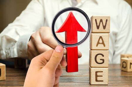 Businessman's hand holds red arrow up near wooden blocks with word Wage. Salary increase concept. Wages rate. Revenue growth and profits. Successful business. Career rise. Promote. Stabilityの写真素材