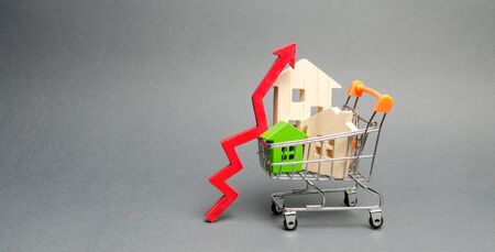 Wooden houses in a shopping cart and red arrow up. The concept of increasing the cost of housing. High demand for real estate. The growth of rent and mortgage rates. Sale of apartmentsの写真素材