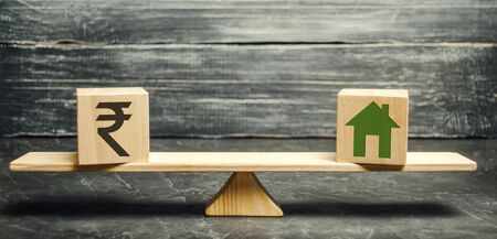 Money - indian rupee (rupiah) and wooden house on scales. Fair value of real estate and housing. Payment of the mortgage interest rates. Redemption of taxes. Tax refund. Apartment valuationの写真素材