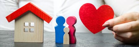 A woman holds in her hands a red heart near a miniature family and home. The concept of property insurance and house. Home Security. Life and health insurance. Protection and safety. Agentの写真素材