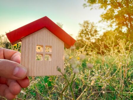 Miniature wooden house in the hands of a man outdoors. Real estate concept. Eco-friendly home. Buying a housing outside the city. The urban downshift. Nature. Fresh air. Mortgage, loan. Red roofの写真素材