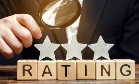 Wooden blocks with the word Rating, three stars and a magnifying glass in the hands of a businessman. The concept of quality of service. Hotel or restaurant rating. Evaluation of critics and visitors.の写真素材